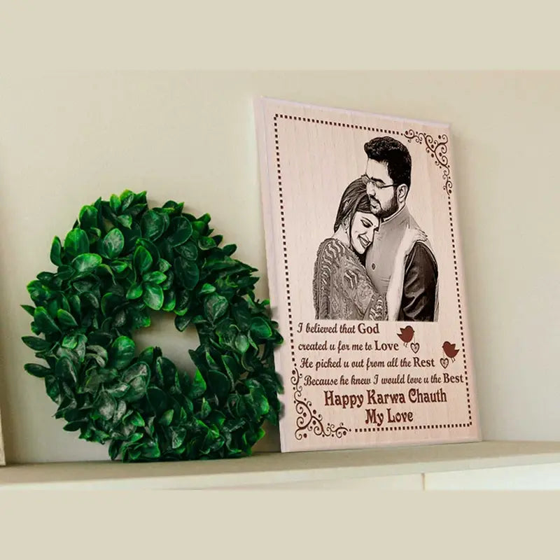 A personalized wooden photo plaque with a printed couple portrait and sentimental text, displayed on a shelf next to a green artificial wreath against a light beige wall.