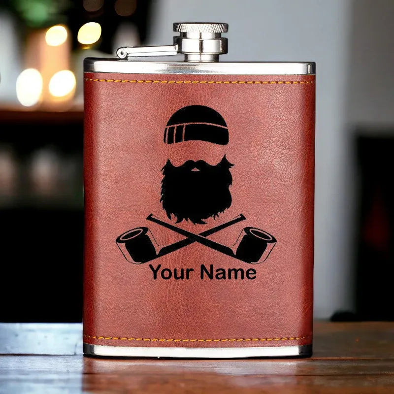 Brown leather flask with a beard and pipes design and 'Your Name' text on a wooden surface.