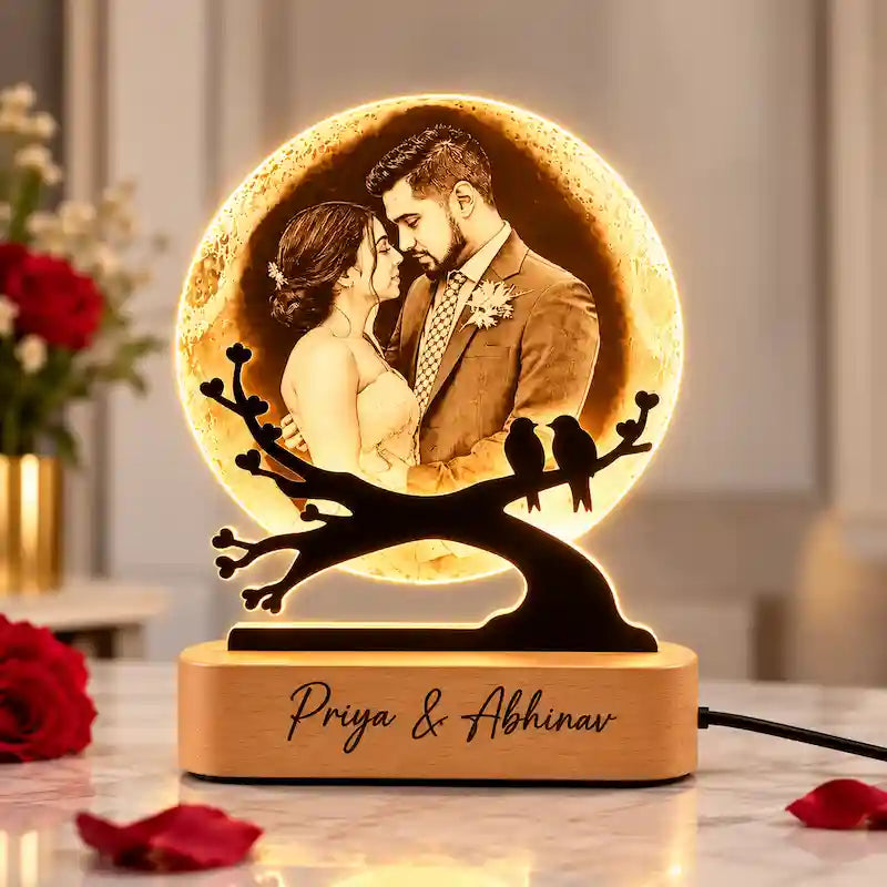Customized 3d moon lamp​ light shaped like a glowing moon with a couple embracing, mounted on a black branch silhouette with two birds, on a wooden base engraved with the names 'Priya & Abhinav'.