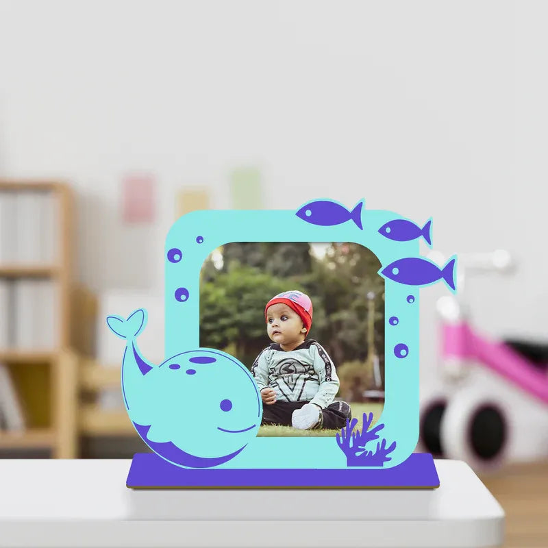 Cute dolphin baby photo frame — teal and blue acrylic standee with whale, fish and coral ocean design, custom baby photo printed inside square opening, purple base, nursery room background.