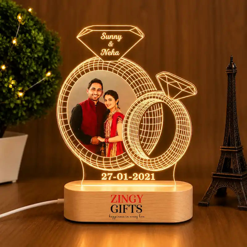 A personalized 3D acrylic LED ring light photo lamp on a wooden base, featuring laser-engraved wedding rings and a diamond shape with custom names 'Sunny & Neha' and the date '27-01-2021'. The base is printed with 'ZINGY GIFTS happiness in every box'.