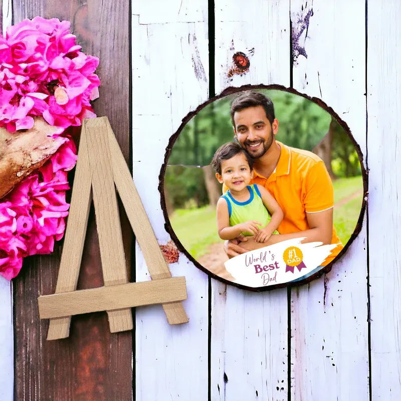 Custom wooden slice photo frame with natural bark edges placed on a small wooden stand, featuring a father and child photo with “World’s Best Dad” text printed at the bottom. The frame is displayed indoors on a table with flowers and a blurred background.