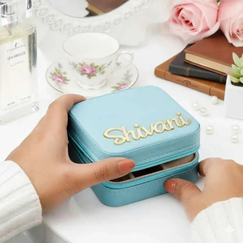 A light blue faux-leather jewelry box with the name 'Shivani' in gold script on the lid, held by two hands over a styled desk with a floral teacup, perfume bottle, books, and pink roses.