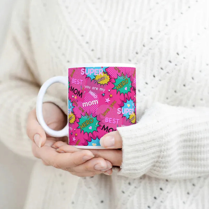Person holding a pink mug with colorful text and graphics, wearing a white sweater.