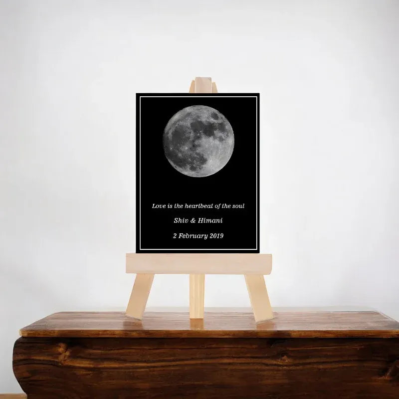 Framed photograph of a moon with personalized text on a wooden stand against a white wall.