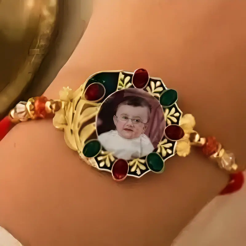 A close-up of a gold-toned traditional bracelet worn on a wrist, featuring a central oval photo frame with red and green stones and red cord accents.