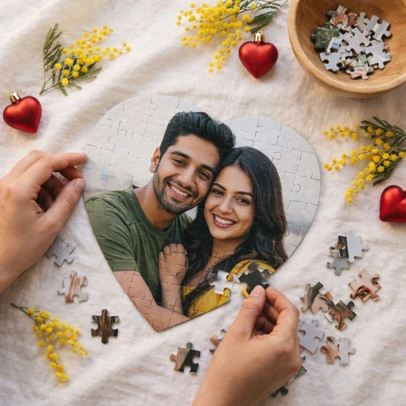 Two people assembling a heart-shaped puzzle with a photo of a couple on a white blanket with decorative elements.