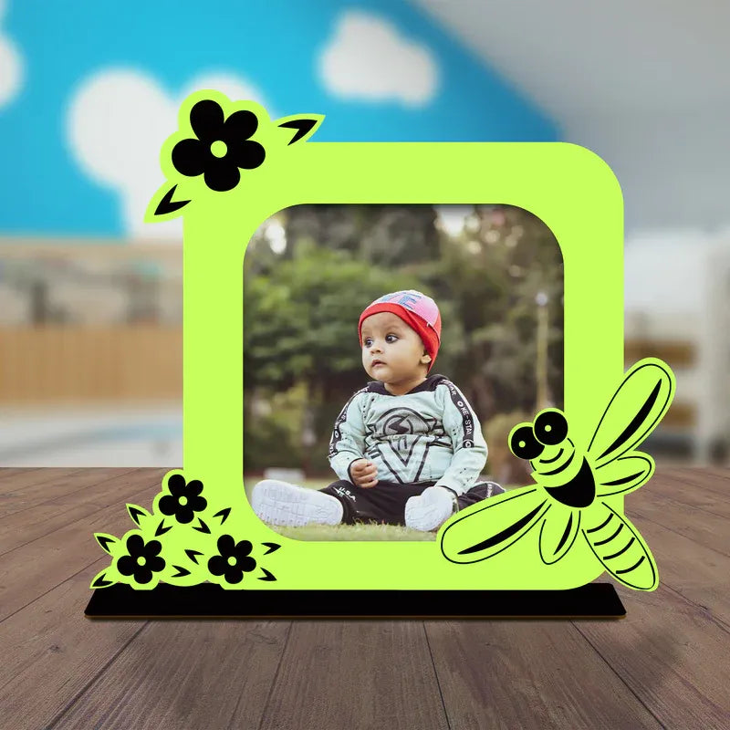 Cute baby photo frame gift — vibrant neon green acrylic frame with bee and flower illustration, personalised baby photo display, solid black stand, nursery room background.