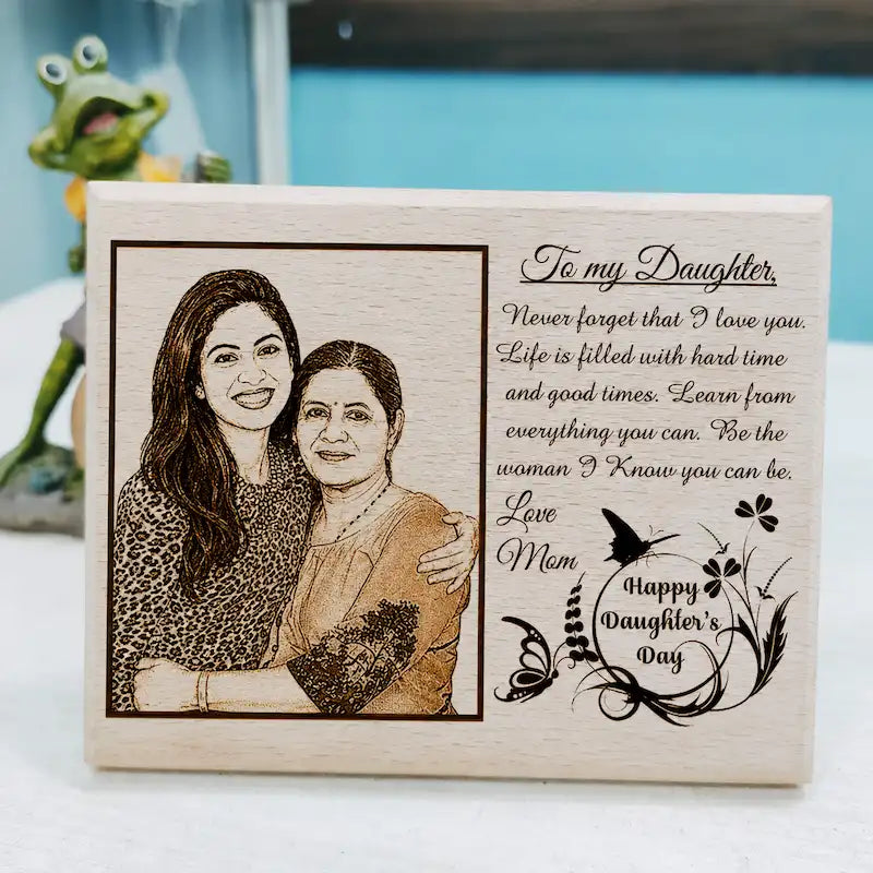 A rectangular wooden engraved gift block for Daughter's Day, featuring a custom photo of two women and a sentimental message for 'my Daughter'. The block is displayed on a white surface with a blurred frog figurine and a blue background.