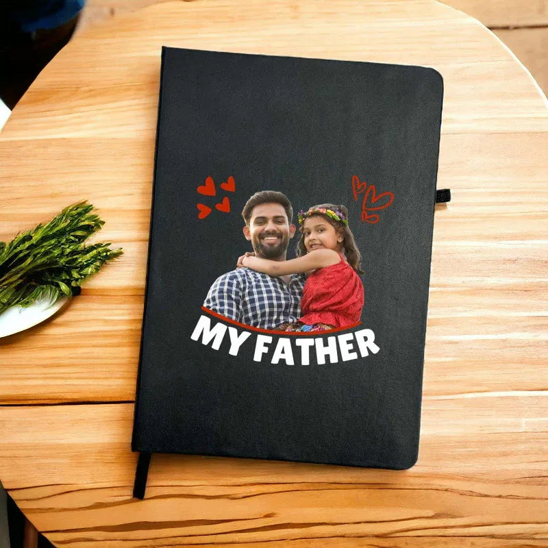 Black notebook with a photo of a man and a child labeled 'MY FATHER' on a wooden table.