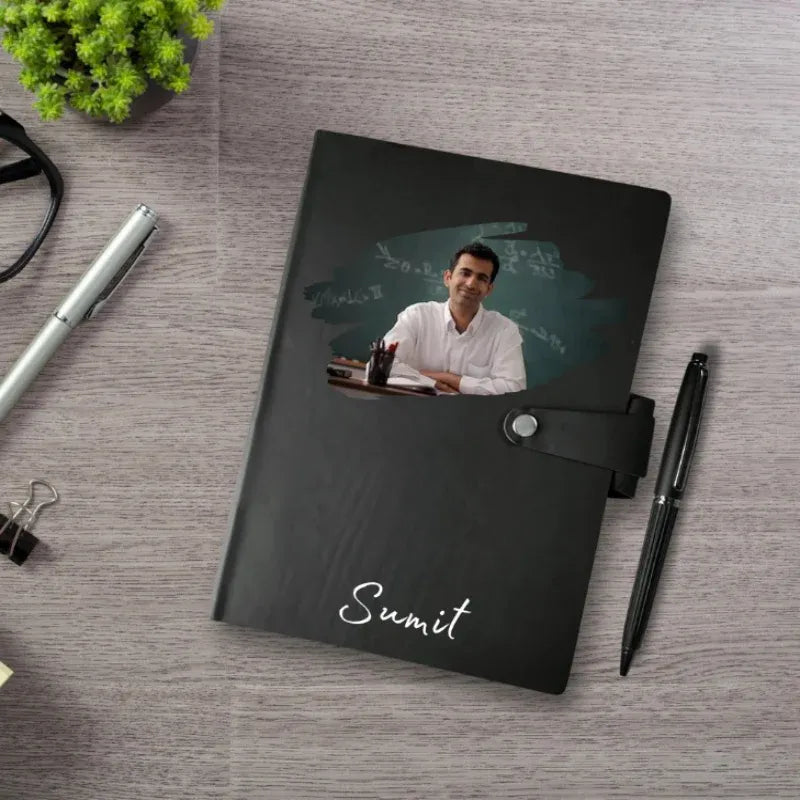 Black notebook with a photo of a man on the cover, placed on a wooden surface with office supplies.