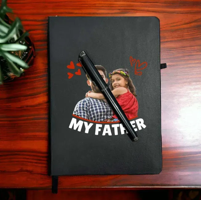Black notebook with a photo of a man and a child, text 'MY FATHER', and a pen on a wooden surface.