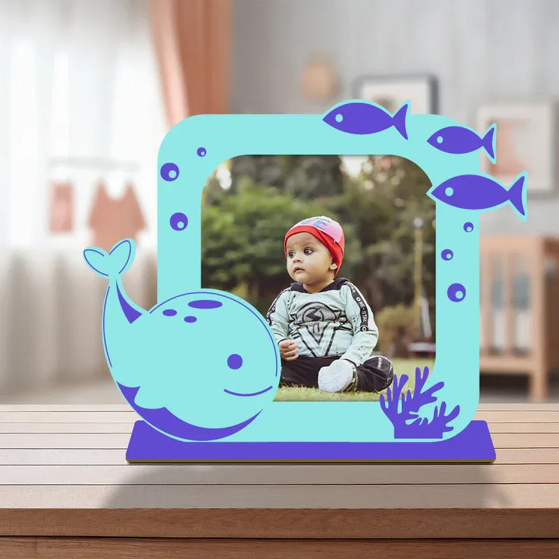 Cute dolphin baby photo frame — vibrant teal acrylic ocean themed standee with smiling whale cartoon, fish illustrations and coral design, personalised baby photo display, purple solid base, desk display.