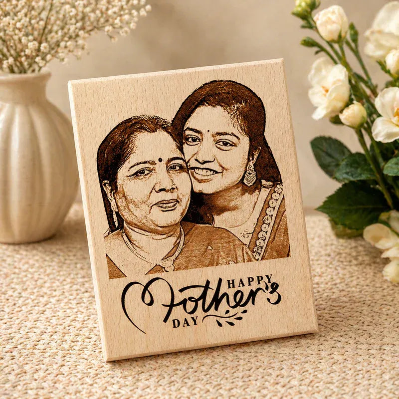 Wooden plaque with engraved portrait of two women and 'Happy Mother's Day' text, placed on a textured surface with flowers in the background.