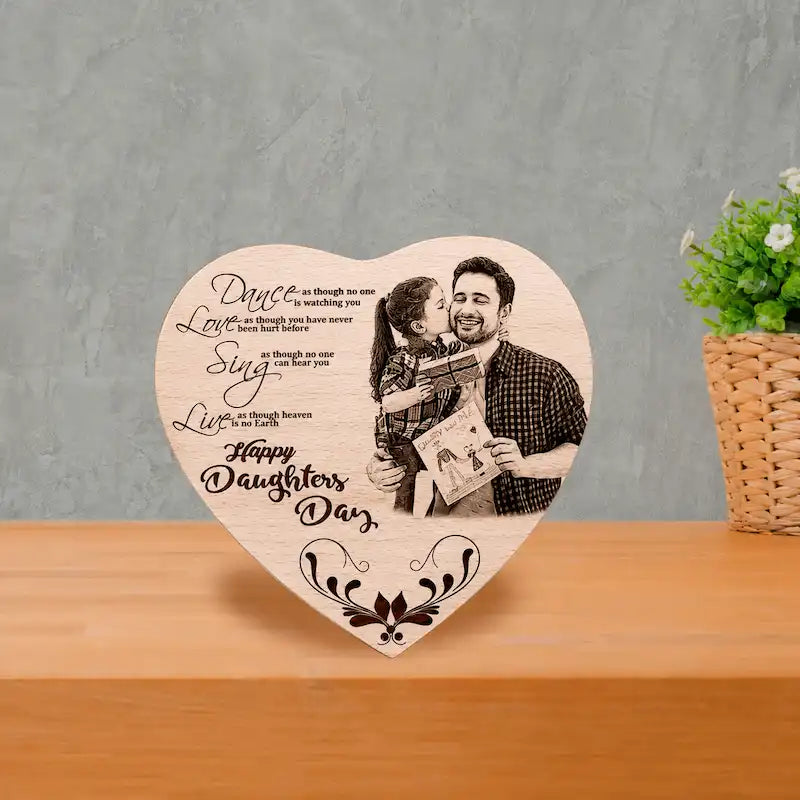 A heart-shaped wooden plaque engraved with father-daughter gifts with a photo of a father and daughter and a 'Happy Daughter's Day' message, displayed on a wooden surface against a gray textured wall.