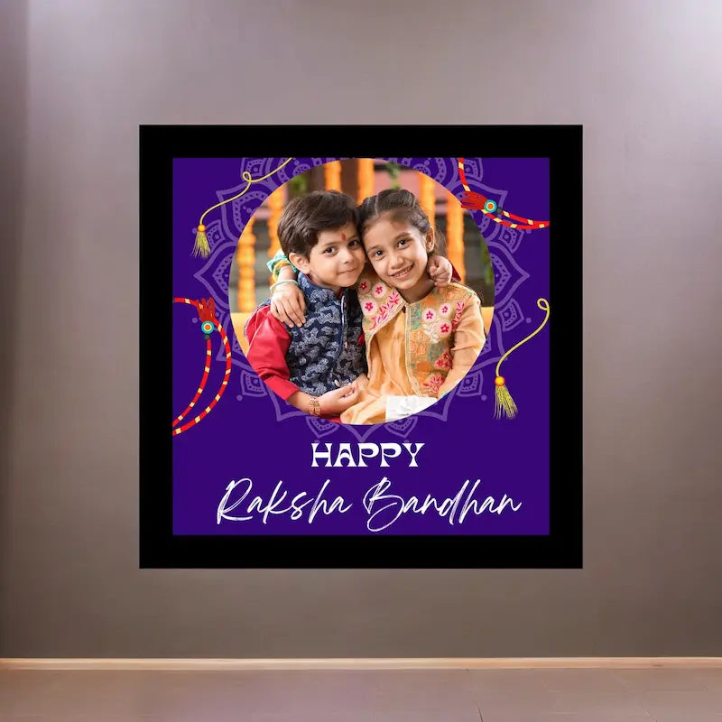 A framed custom photo print with a purple festive background and the text 'HAPPY Raksha Bandhan' is displayed on a gray wall. The print features a circular photo of two children, traditional Indian decorative motifs, and a black frame.