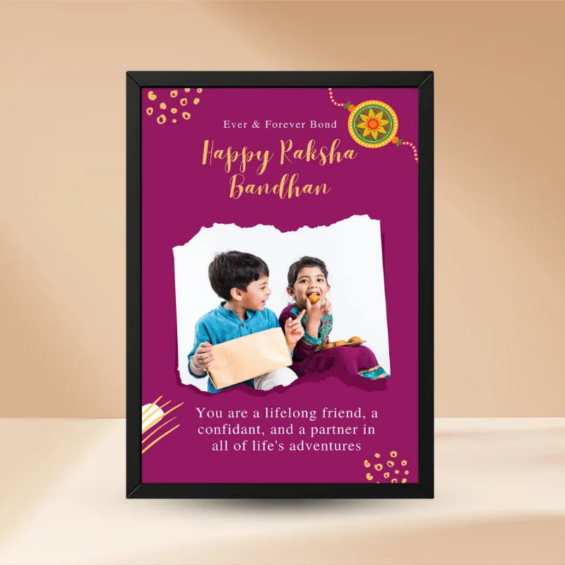 A framed personalized Raksha Bandhan greeting card or print with a black frame, set against a warm beige background. The card features a deep magenta background with gold and white text reading 'Happy Raksha Bandhan' and 'Ever & Forever Bond,' along with decorative motifs and a photo of two children.