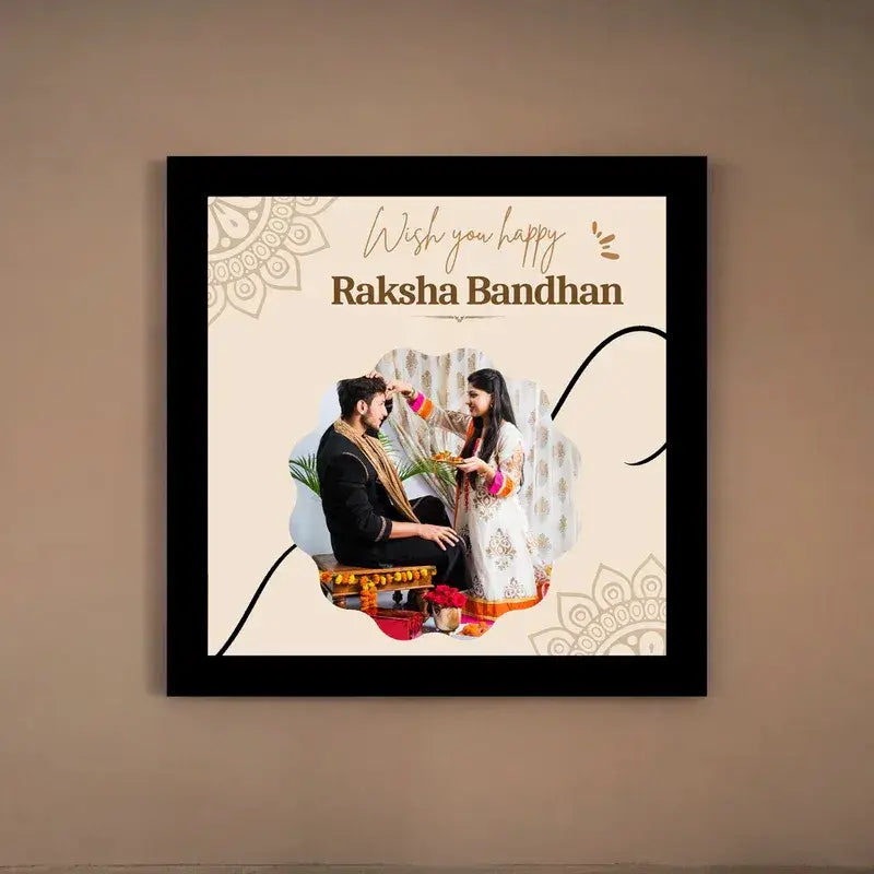 A framed square wall art print hanging on a beige wall. The print features the text 'Wish you happy Raksha Bandhan' above a photo of a couple in traditional Indian attire, with decorative mandala-style motifs and a black frame.