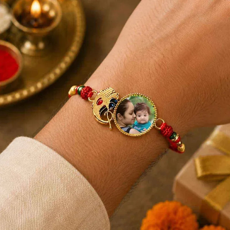 A close-up of a person's wrist wearing a decorative gold-toned Rakhi bracelet with red and green thread beads and a custom photo charm showing two children.