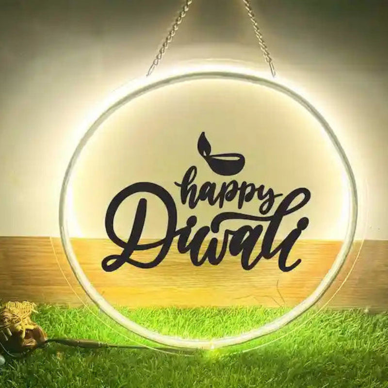 A glowing round LED sign hanging by chains, featuring the text 'happy Diwali' in black script with a small diya icon above it. The sign is shown illuminated against a warm background with decorative grass and small ornaments at the bottom.
