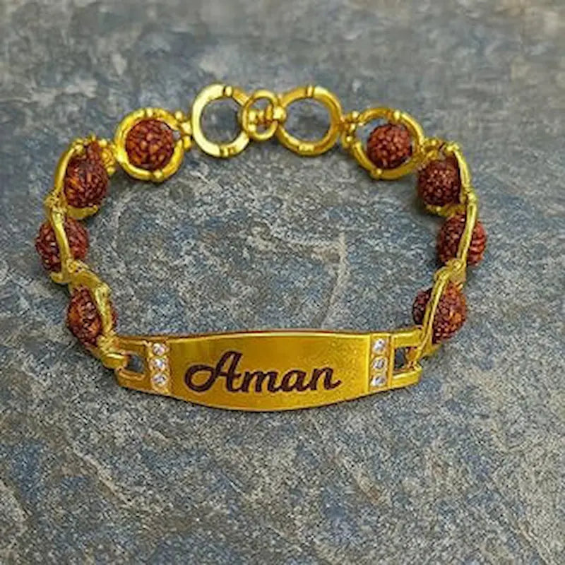 A gold-plated ID bracelet with a personalized nameplate engraved with 'Aman' and small clear rhinestones, featuring textured reddish-brown rudraksha beads separated by gold rings, displayed on a gray stone surface.