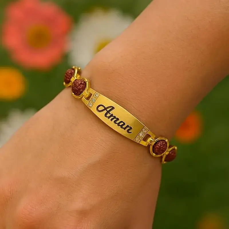A gold-plated ID bracelet with the name 'Aman' engraved in black script, decorated with red rudraksha beads and small clear crystals, shown worn on a person's wrist against a blurred green and orange background.