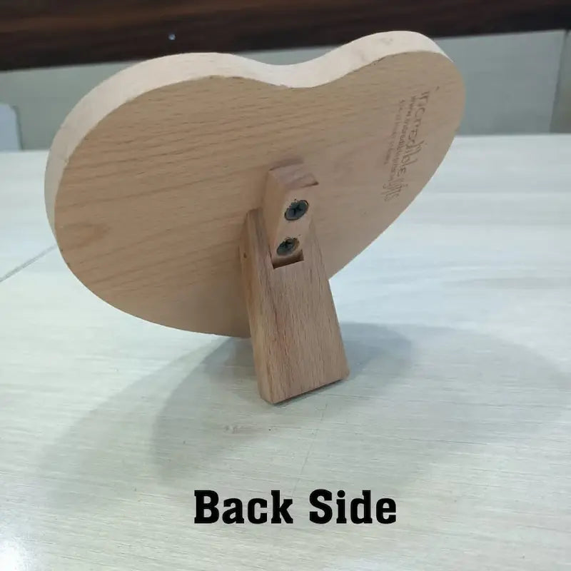A heart-shaped wooden picture frame shown from the back, resting on a light-colored tabletop. The image displays the natural wood grain backplate and the mounting bracket with two visible screws.