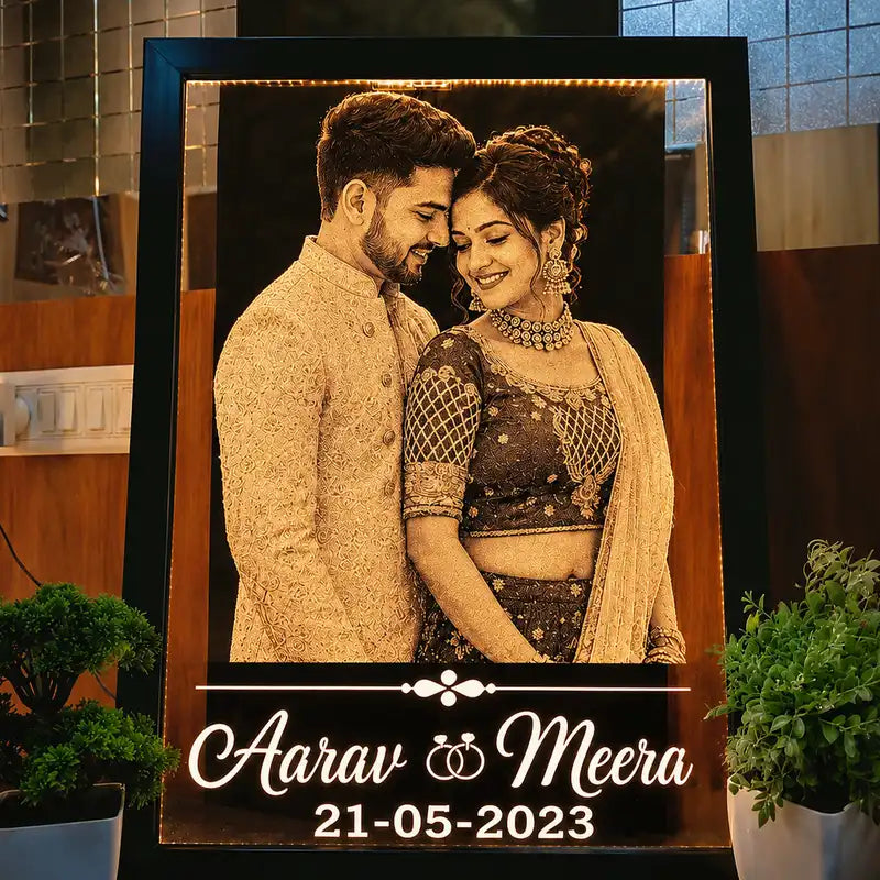 A custom acrylic led photo frame featuring a couple in traditional Indian attire, with the names 'Aarav & Meera' and the date '21-05-2023' printed at the bottom. The framed artwork is displayed on a shelf in a home interior setting.