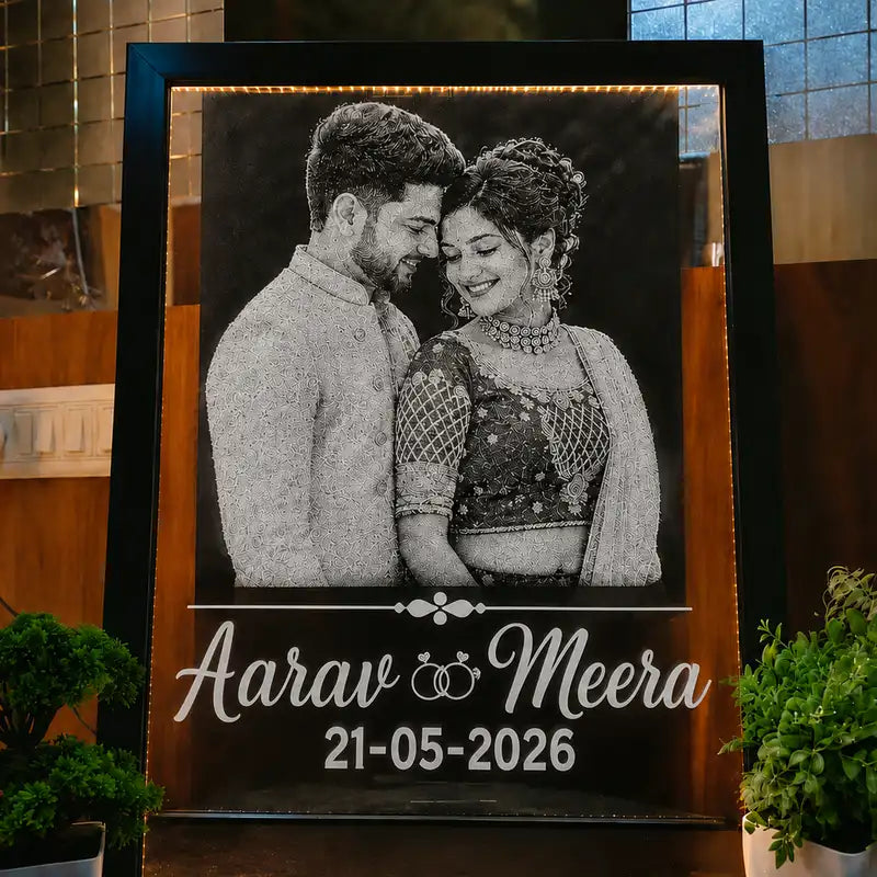 A custom framed wedding photo sign featuring a black-and-white portrait of a couple with the names 'Aarav Meera' and the date '21-05-2026' in white script and numerals on a dark background, displayed indoors with plants around it.