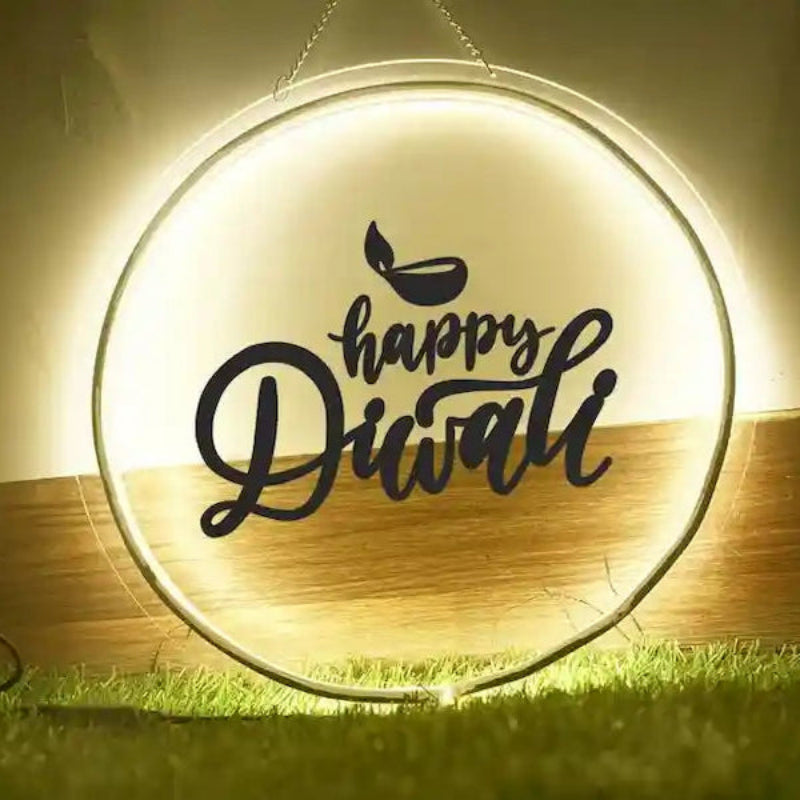 A circular LED light sign with a glowing warm white background and black script text reading 'Happy Diwali' with a small diya icon above, shown hanging above grass against an indoor wall.