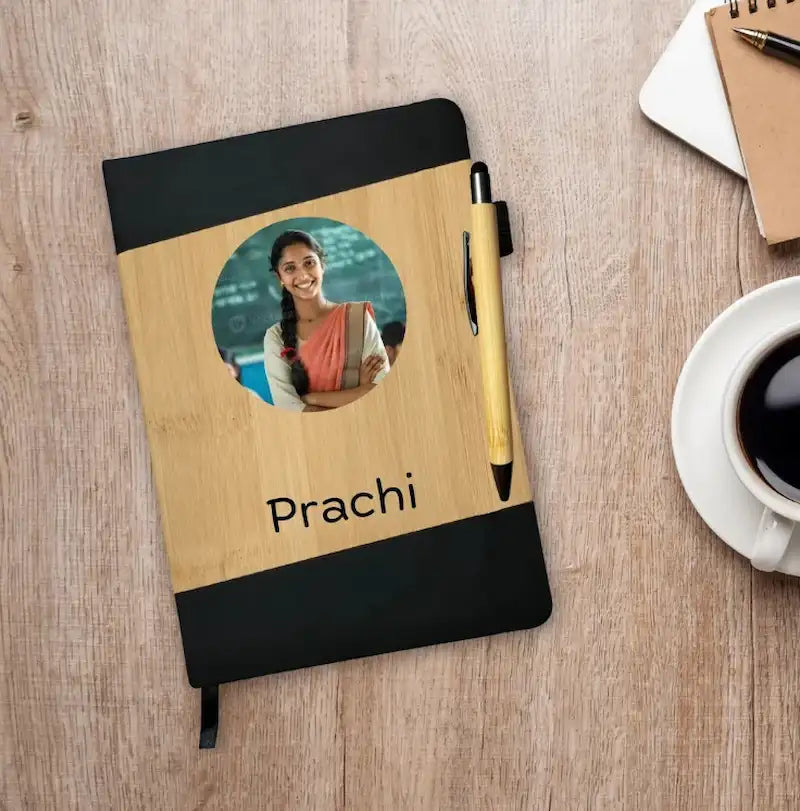 A personalized bamboo notebook with black covers on the top and bottom, featuring a circular photo cutout of a birthday gift for teacher​ woman and the name 'Prachi' printed in black. A matching wooden pen is tucked into the notebook, which is displayed on a light wood desk alongside a coffee cup, another notebook, and a pen.