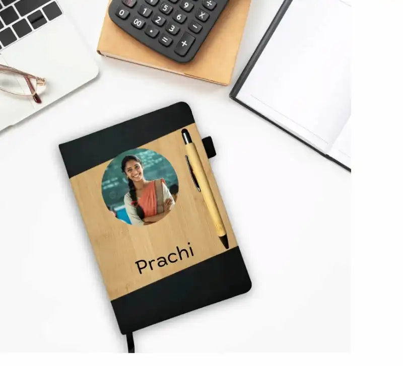 A black and light wood notebook with a custom photo of a woman and the name 'Prachi' printed on the front, styled on a white desk with a pen, glasses, a laptop, and a calculator.