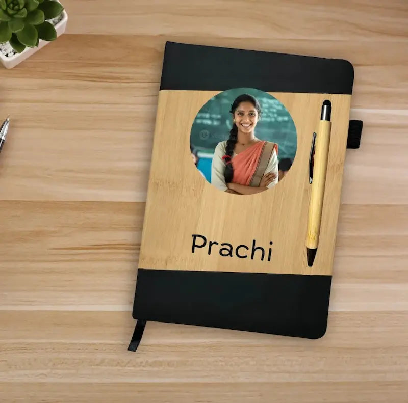 A personalized hardcover notebook with a light wood grain center panel, black top and bottom borders, a circular photo cutout, the name 'Prachi' printed below, and a pen attached on a wooden desk.