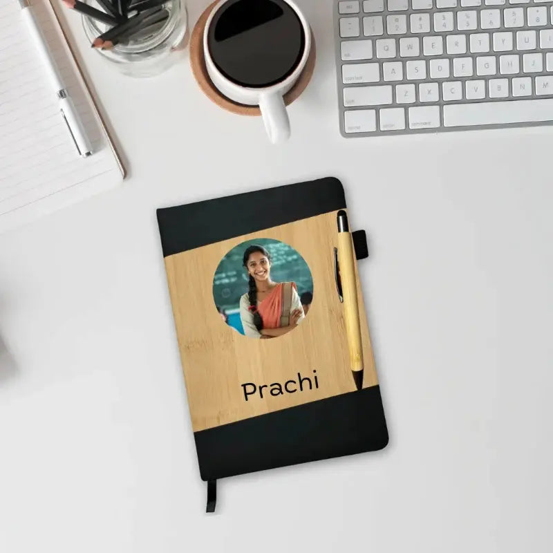 A personalized notebook with a black and light wood cover, featuring a circular photo cutout of a woman, the name 'Prachi', and a gold pen, shown on a white desk with a keyboard, coffee mug, and other office items.