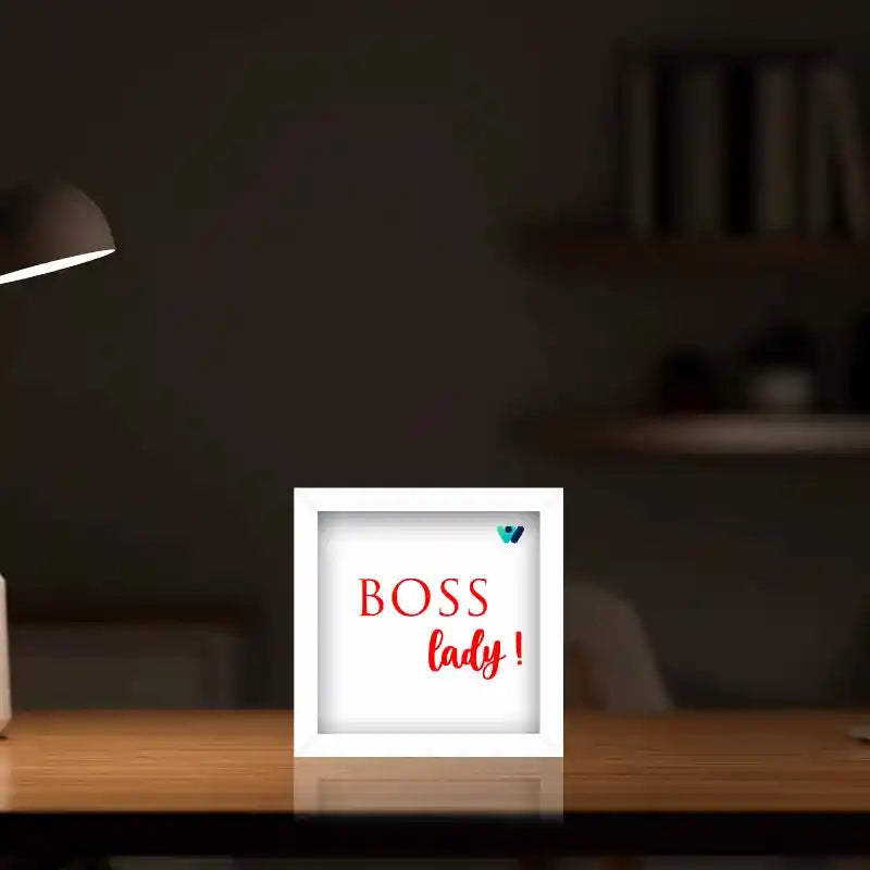 A square white desk lamp sign with the text 'BOSS lady!' in red, placed on a wooden desk in a dimly lit room with a blurred lamp in the background.