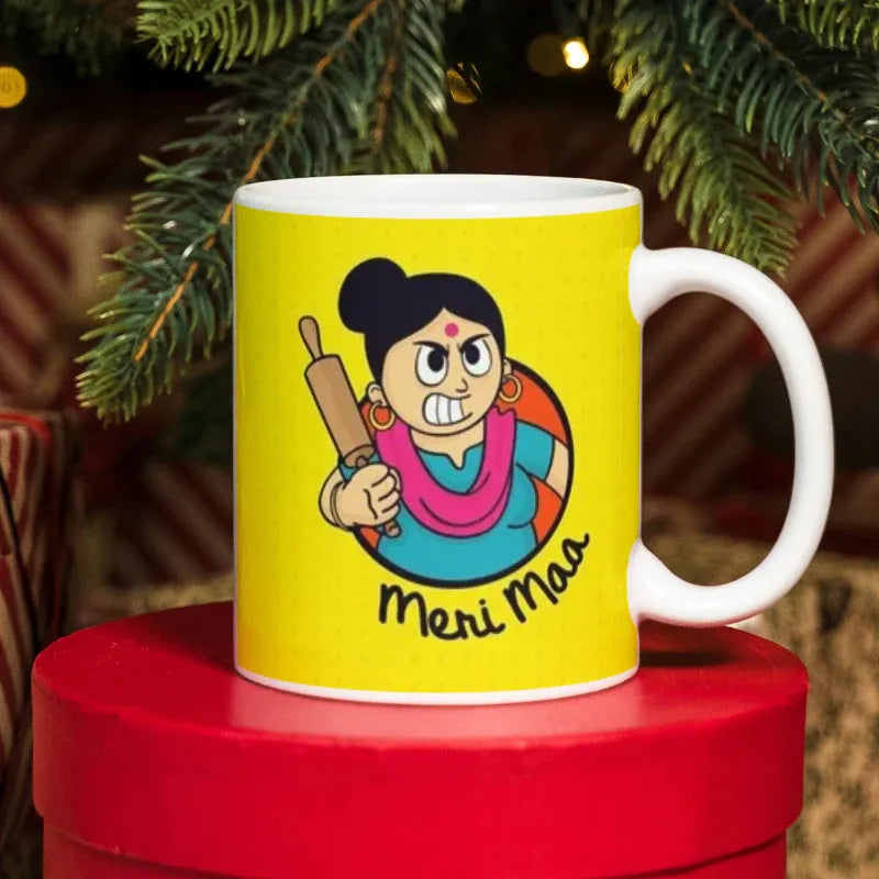 Yellow mug with a cartoon character and 'Meri Maa' text on a red surface with Christmas tree background