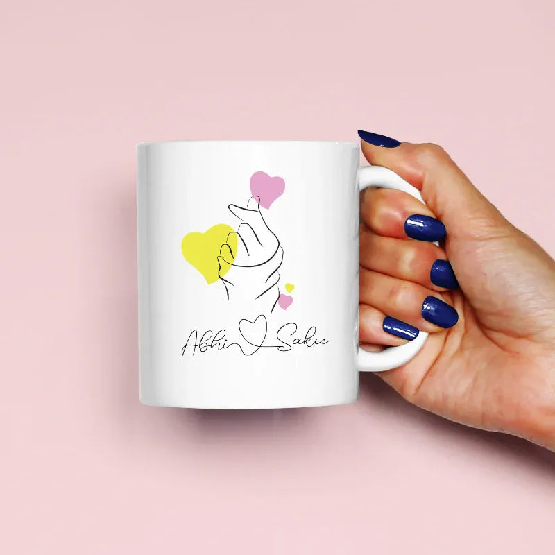Hand holding a mug with a design of two hands holding hearts and the text 'Abhi & Saki' on a pink background.