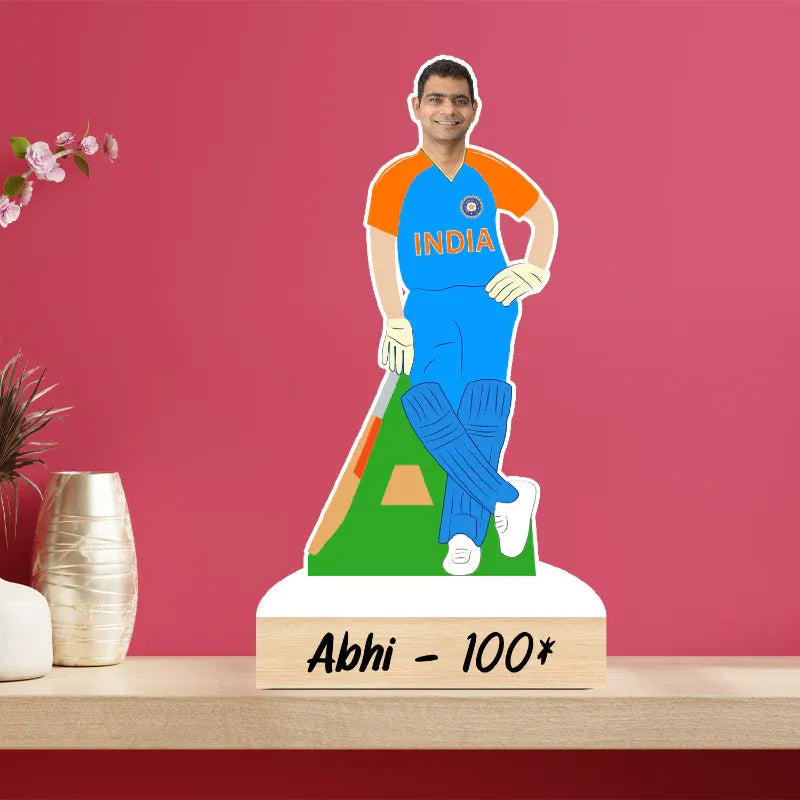Ceramic figure of a cricketer in an India jersey on a wooden base with 'Abhi - 100*' text, against a pink background.