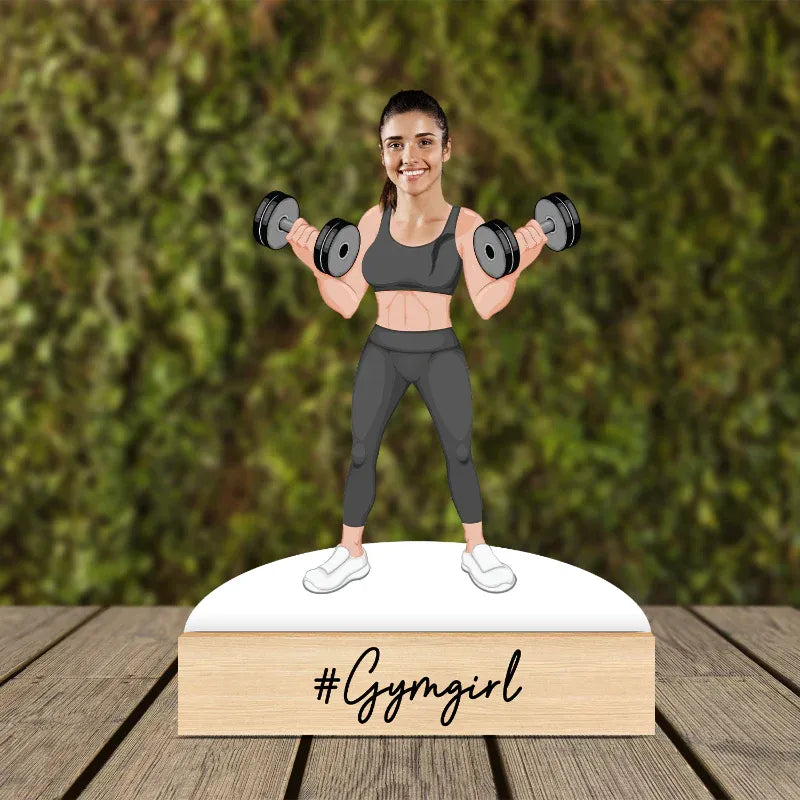 
Figurine of a woman lifting weights with '#Gymgirl' text on a wooden base against a green foliage 