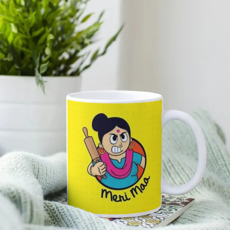 Yellow mug with a cartoon character holding a rolling pin and text 'Meri Maa' on a white surface.