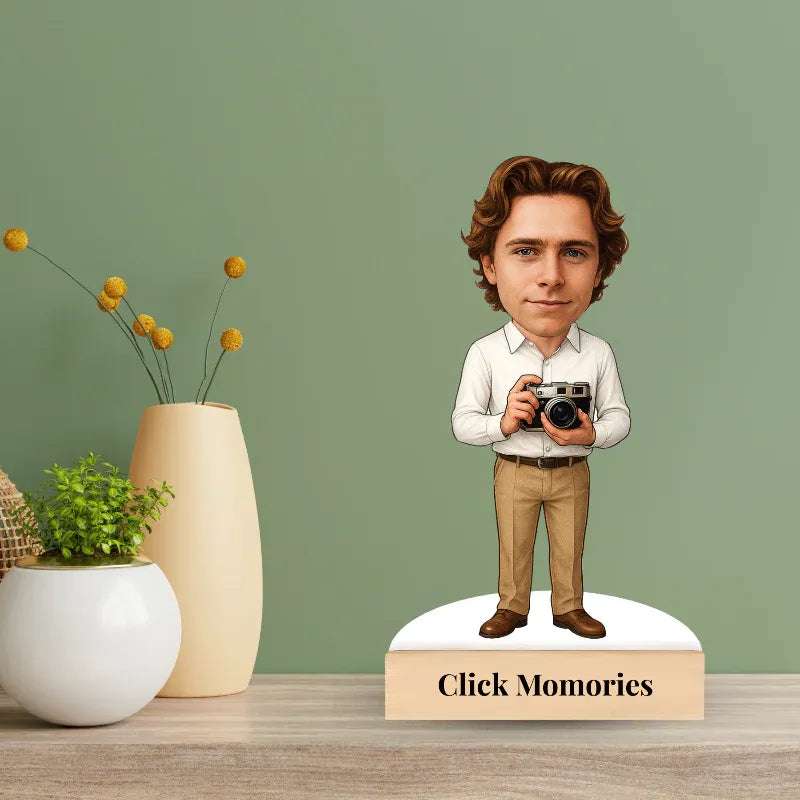 Figurine of a person holding a camera on a stand with 'Click Memories' branding, against a green wall with decorative items.