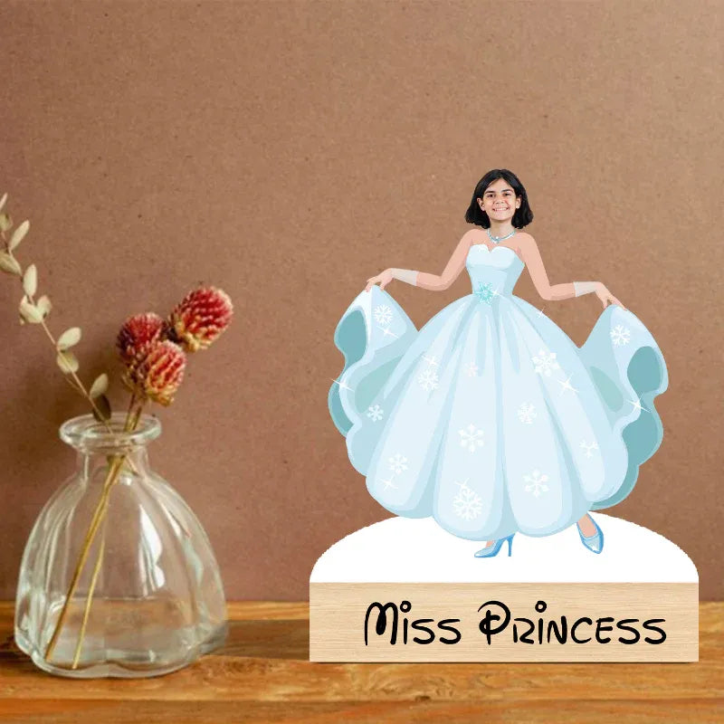 Figurine of a woman in a blue dress labeled 'Miss Princess' next to a vase with flowers on a wooden surface.