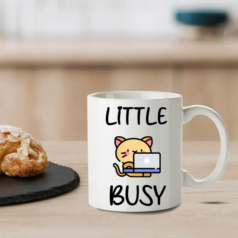 White mug with a cat illustration and text 'LITTLE BUSY' on a wooden surface with a pastry.