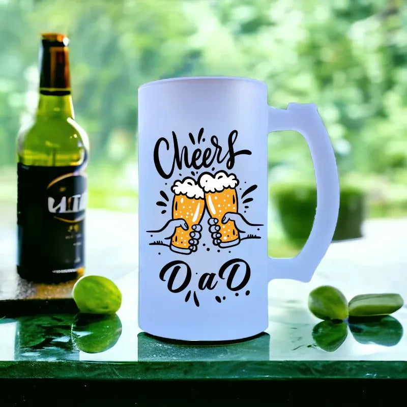 White mug with 'Cheers DAD' text and beer graphics next to a bottle on a green surface.