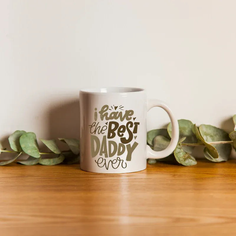 White mug with 'I have the best daddy ever' text on a wooden surface with eucalyptus leaves.
