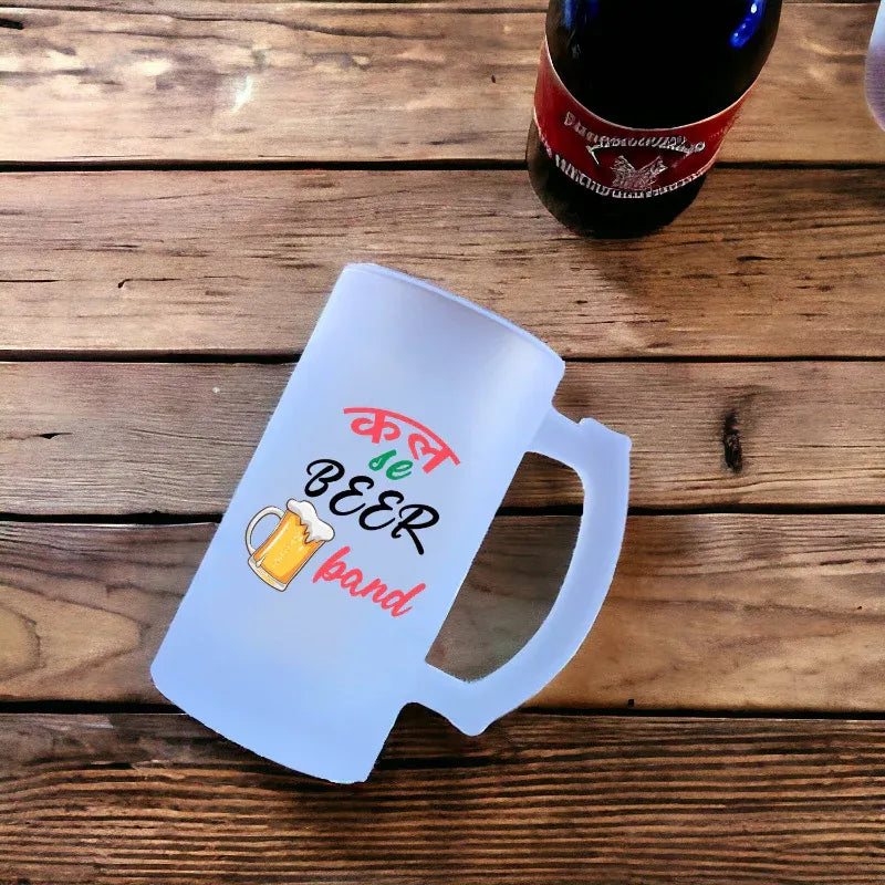 Frosted mug with colorful text and a beer graphic on a wooden surface next to a bottle.
