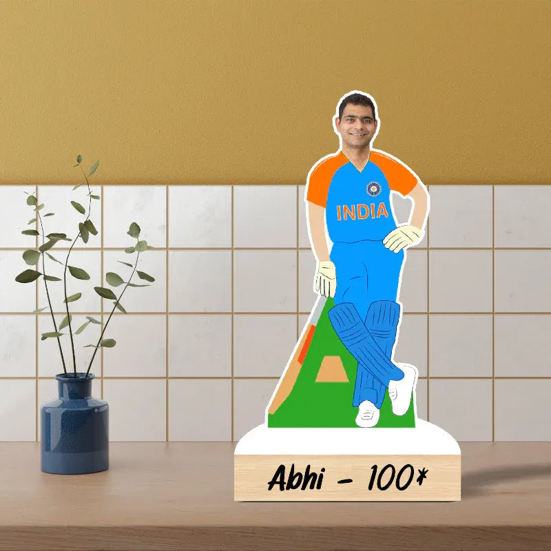 Cardboard cutout of a person in an India sports uniform on a stand with 'Abhi - 100*' text, against a tiled wall background.