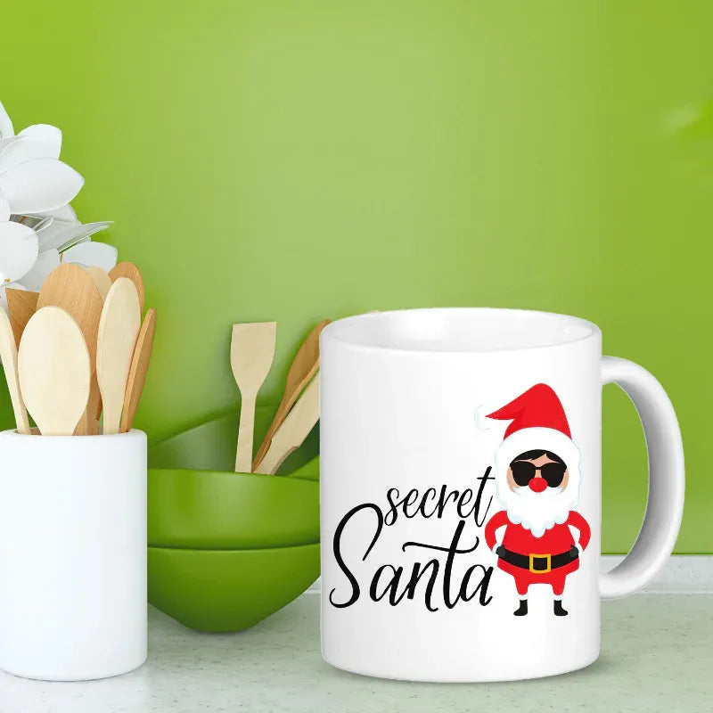 White mug with 'secret Santa' design next to kitchen utensils on a green background