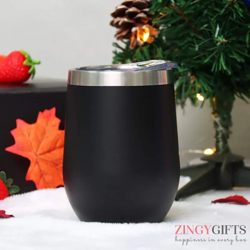 Black insulated tumbler with silver lid on a snowy surface with Christmas decorations.