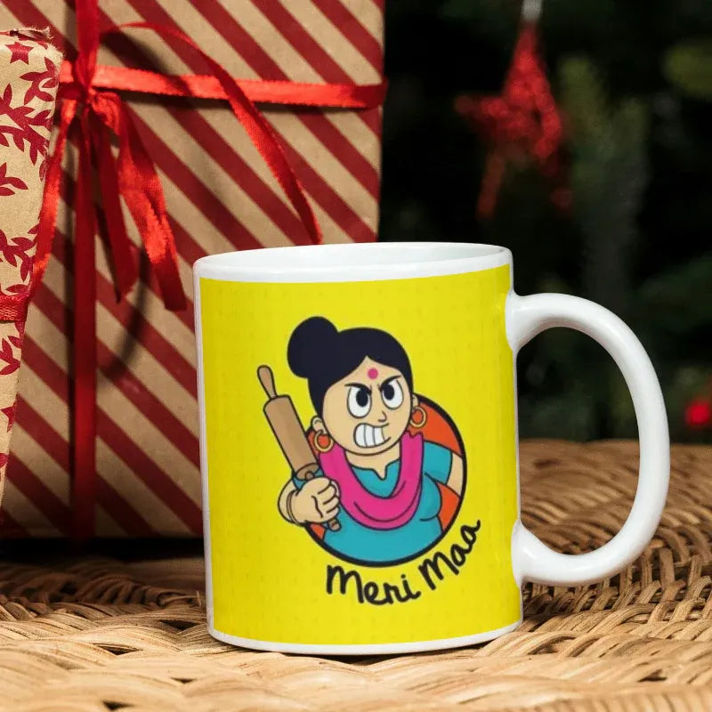 Yellow mug with cartoon character and 'Meri Mas' text, placed on a woven surface with a gift box in the background.